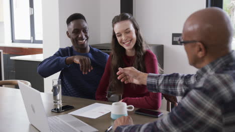 Happy-diverse-couple-and-male-financial-advisor-using-laptop-shaking-hands-at-home,slow-motion