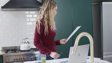 Biracial-woman-using-laptop-and-holding-documents-in-kitchen-at-home,-slow-motion
