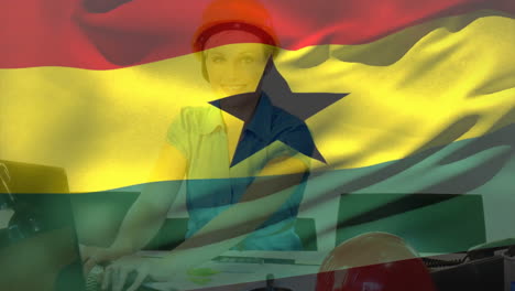 Animation-of-ghana-flag-waving-over-smiling-caucasian-woman-wearing-helmet-working-on-computer