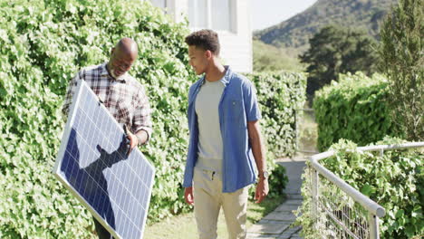 Padre-Afroamericano-Sosteniendo-Panel-Solar-Hablando-Con-Un-Hijo-Adulto-En-Un-Jardín-Soleado,-Cámara-Lenta
