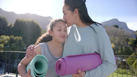 Happy-biracial-mother-and-daughter-standing-with-yoga-mats-on-terrace-in-sunny-day,-slow-motion