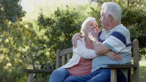 Happy-senior-caucasian-couple-sitting-on-bench-embracing-in-sunny-garden,-slow-motion