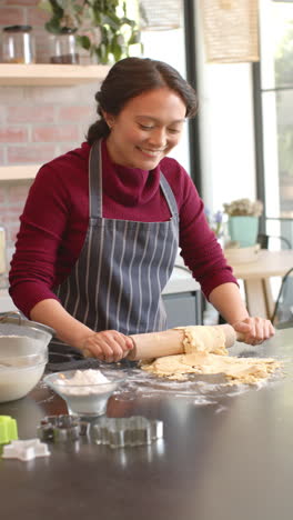 Vertical-video-of-happy-biracial-woman-in-apron-rolling-dough-in-kitchen-at-home,-slow-motion