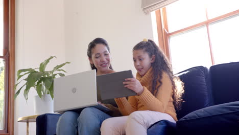 Happy-biracial-mother-and-daughter-sitting-on-sofa,-using-laptop-and-tablet-in-sunny-living-room