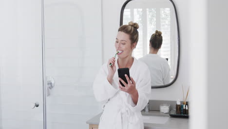 Glückliche-Biracial-Frau-Putzt-Zähne-Mit-Smartphone-Im-Badezimmer,-Zeitlupe