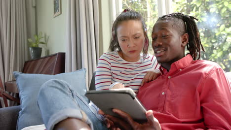 Happy-diverse-couple-sitting-on-couch-using-tablet-in-sunny-living-room,-slow-motion