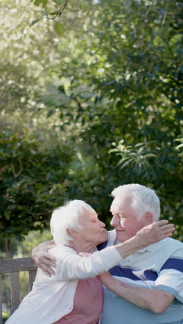 Vertital-video-of-senior-caucasian-couple-sitting-on-bench-embracing-in-sunny-garden,-slow-motion
