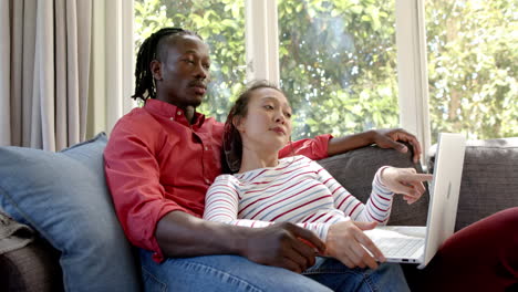 Happy-diverse-couple-relaxing-on-couch-using-laptop-in-sunny-living-room,-slow-motion