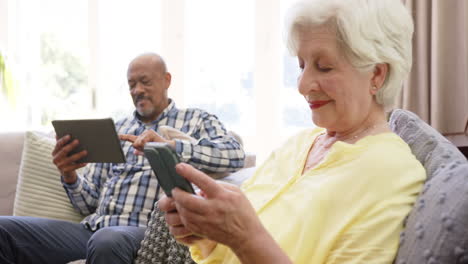 Happy-diverse-senior-couple-using-smartphone-and-tablet-in-sunny-living-room,-slow-motion