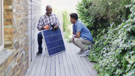 Padre-Afroamericano-Mostrando-Panel-Solar-A-Hijo-Adulto-En-Jardín-Soleado,-Espacio-Para-Copiar,-Cámara-Lenta