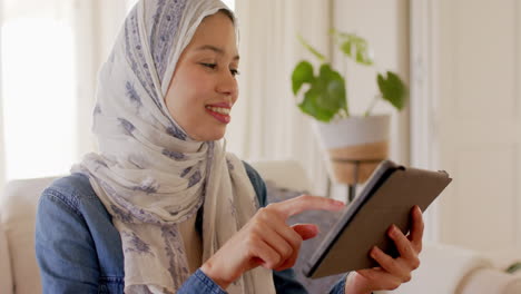 Happy-biracial-woman-in-hijab-using-tablet-on-sofa-at-home-with-copy-space,-slow-motion