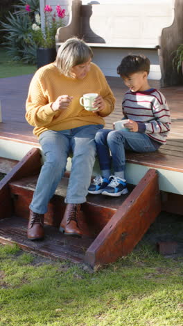 Vertical-video-of-biracial-grandmother-and-grandson-sitting-on-terrace-and-holding-mugs,-slow-motion