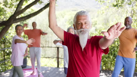 Diverse-senior-male-instructor-and-friends-practicing-yoga-standing-in-sunny-nature,-slow-motion