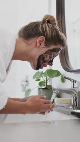 Vertical-video-of-biracial-woman-washing-beauty-face-mask-in-bathroom,-slow-motion