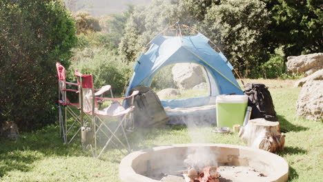 Smoking-firepit,-tent,-chairs-and-camping-equipment-in-sunny-countryside,-copy-space,-slow-motion