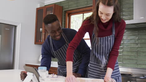 Happy-diverse-couple-standing-in-kitchen,-using-tablet-and-rolling-out-dough,slow-motion