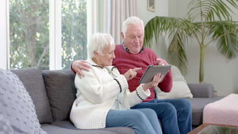 Happy-senior-caucasian-couple-sitting-on-sofa-using-tablet-at-home,-slow-motion
