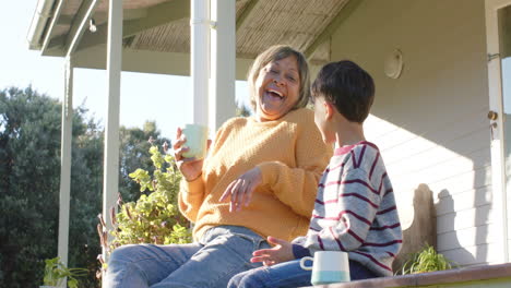 Happy-senior-biracial-grandmother-and-grandson-holding-mugs-sitting-on-terrace,-slow-motion