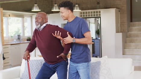 Glücklicher-Afroamerikanischer-Erwachsener-Sohn-Hilft-älterem-Vater,-Zu-Hause-Von-Der-Couch-Aufzustehen,-Zeitlupe