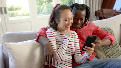 Happy-diverse-couple-sitting-on-couch-using-smartphone-in-sunny-living-room,-slow-motion