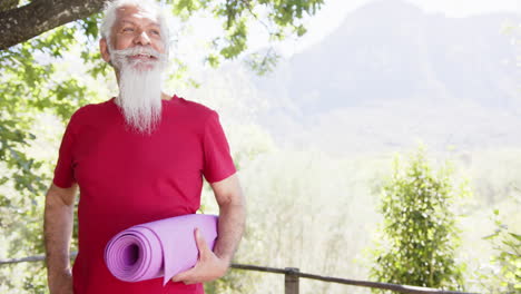 Happy-biracial-senior-man-with-white-beard-holding-yoga-mat-smiling-in-sun,-copy-space,-slow-motion
