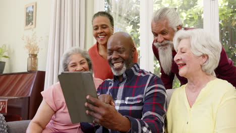 Happy-diverse-senior-male-and-female-friends-using-tablet-together-in-sunny-living-room,-slow-motion