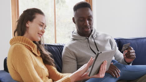 Happy-diverse-couple-sitting-on-sofa,-using-tablet-and-credit-card-in-home,copy-space