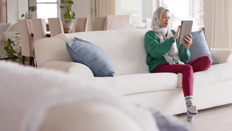 Mujer-Birracial-En-Hijab-Usando-Tableta-En-El-Sofá-En-Casa-Con-Espacio-Para-Copiar,-Cámara-Lenta