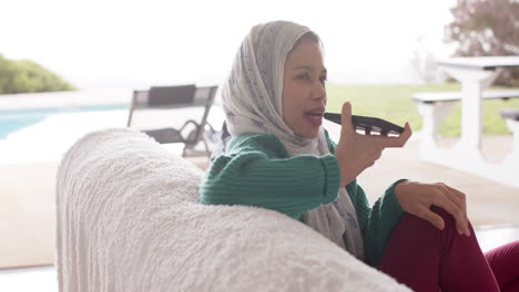 Biracial-woman-in-hijab-using-smartphone-on-sofa-at-home-with-copy-space,-slow-motion