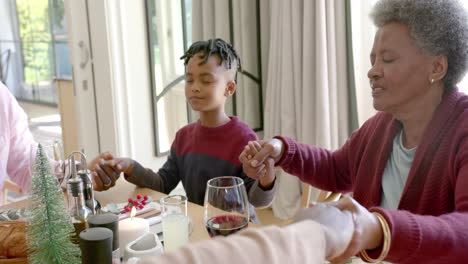Happy-multi-generation-african-american-family-holding-hands-saying-grace-at-christmas-dinner-table