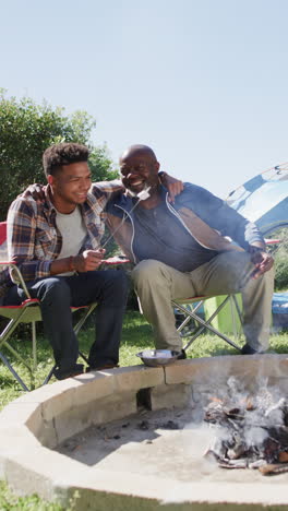Vertical-video-of-happy-african-american-father-and-son-cooking-on-campfire-in-the-sun,-slow-motion