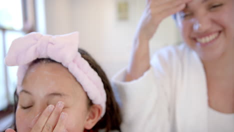 Glückliche-Biracial-Mutter-Und-Tochter-Mit-Stirnbändern-Creme-Auf-Gesichter-In-Sonnigen-Badezimmer