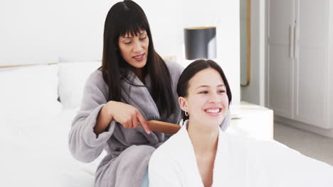 Happy-biracial-mother-in-bathrobe-brushing-hair-of-smiling-adult-daughter-in-sunny-room,-slow-motion