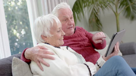 Happy-senior-caucasian-couple-sitting-on-sofa-using-tablet-at-home,-slow-motion