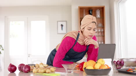 Mujer-Birracial-Con-Hijab-Usando-Tableta-Preparando-Comida-En-La-Cocina-De-Casa-Con-Espacio-Para-Copiar,-Cámara-Lenta