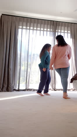 Vertical-video-of-happy-biracial-mother-and-daughter-dancing-and-smiling-in-sunny-bedroom