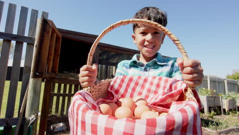 Portrait-of-happy-biracial-boy-holding-basket-with-eggs-next-to-henhouse,-slow-motion