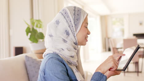 Feliz-Mujer-Birracial-En-Hijab-Usando-Tableta-En-El-Sofá-En-Casa-Con-Espacio-Para-Copiar,-Cámara-Lenta