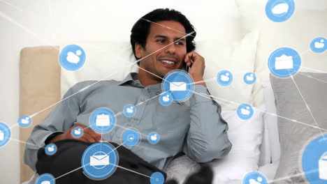 Animation-of-connected-icons-over-smiling-biracial-man-sitting-on-sofa-and-talking-on-smartphone