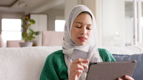 Mujer-Birracial-En-Hijab-Usando-Tableta-En-El-Sofá-En-Casa-Con-Espacio-Para-Copiar,-Cámara-Lenta