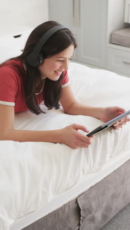 Vertical-video-of-happy-biracial-woman-in-headphones-lying-on-bed-using-tablet,-slow-motion