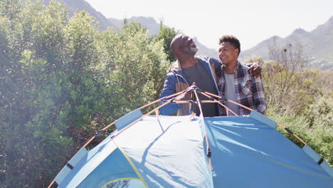 Happy-african-american-father-and-son-pitching-tent-embracing-in-sunny-countryside,-slow-motion