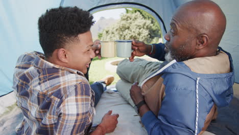 Glücklicher-Afroamerikanischer-Vater-Und-Erwachsener-Sohn-Machen-Einen-Toast-Mit-Kaffeetassen-Im-Zelt,-Zeitlupe