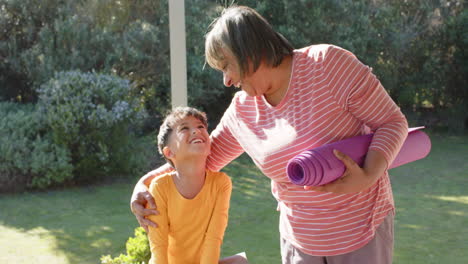 Portrait-of-happy-senior-biracial-grandmother-and-grandson-holding-yoga-mat-on-terrace,-slow-motion