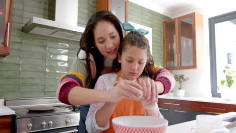 Happy-biracial-mother-and-daughter-breaking-eggs-and-smiling-in-sunny-kitchen