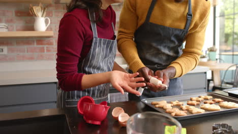 Happy-diverse-couple-in-aprons-decorating-christmas-cookies-in-kitchen,-slow-motion
