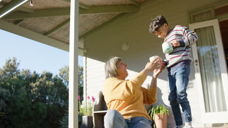 Happy-senior-biracial-grandmother-and-grandson-holding-mugs-on-terrace,-slow-motion
