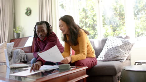 Happy-diverse-couple-with-laptop-discussing-domestic-finance-in-sunny-home,-copy-space,-slow-motion