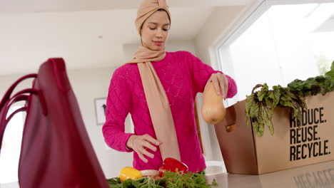 Feliz-Mujer-Birracial-En-Hijab-Con-Compras-En-La-Cocina-De-Casa-Con-Espacio-Para-Copiar,-Cámara-Lenta