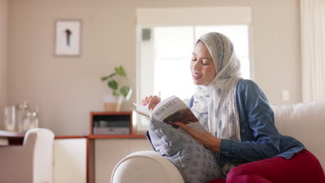 Happy-biracial-woman-in-hijab-reading-book-on-sofa-at-home-with-copy-space,-slow-motion